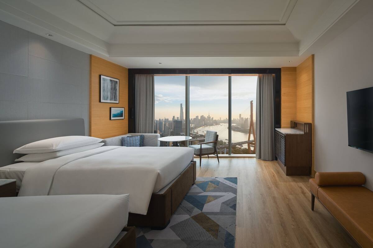 Habitación de hotel moderna con una cama grande, un escritorio, una silla y un televisor. La habitación tiene un ventanal con vistas a la ciudad.