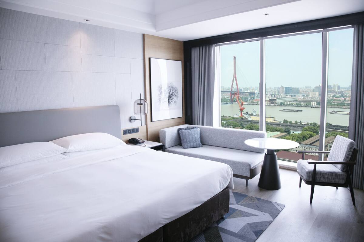 Habitación de hotel moderna con una cama grande, un sofá, una mesita y una silla. Cuenta con un ventanal amplio que ofrece vistas a un paisaje urbano y una grúa.