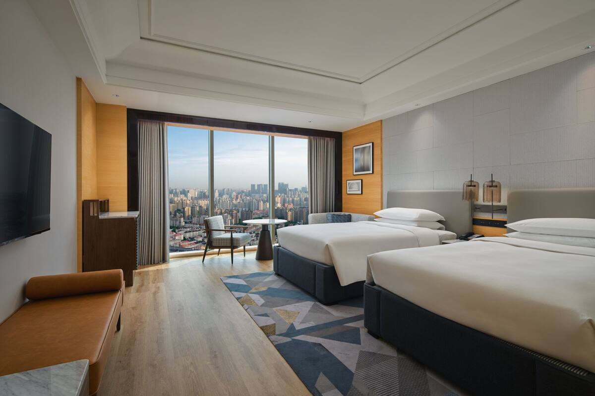 Habitación de hotel con dos camas, un escritorio, una silla y un ventanal con vistas a la ciudad.