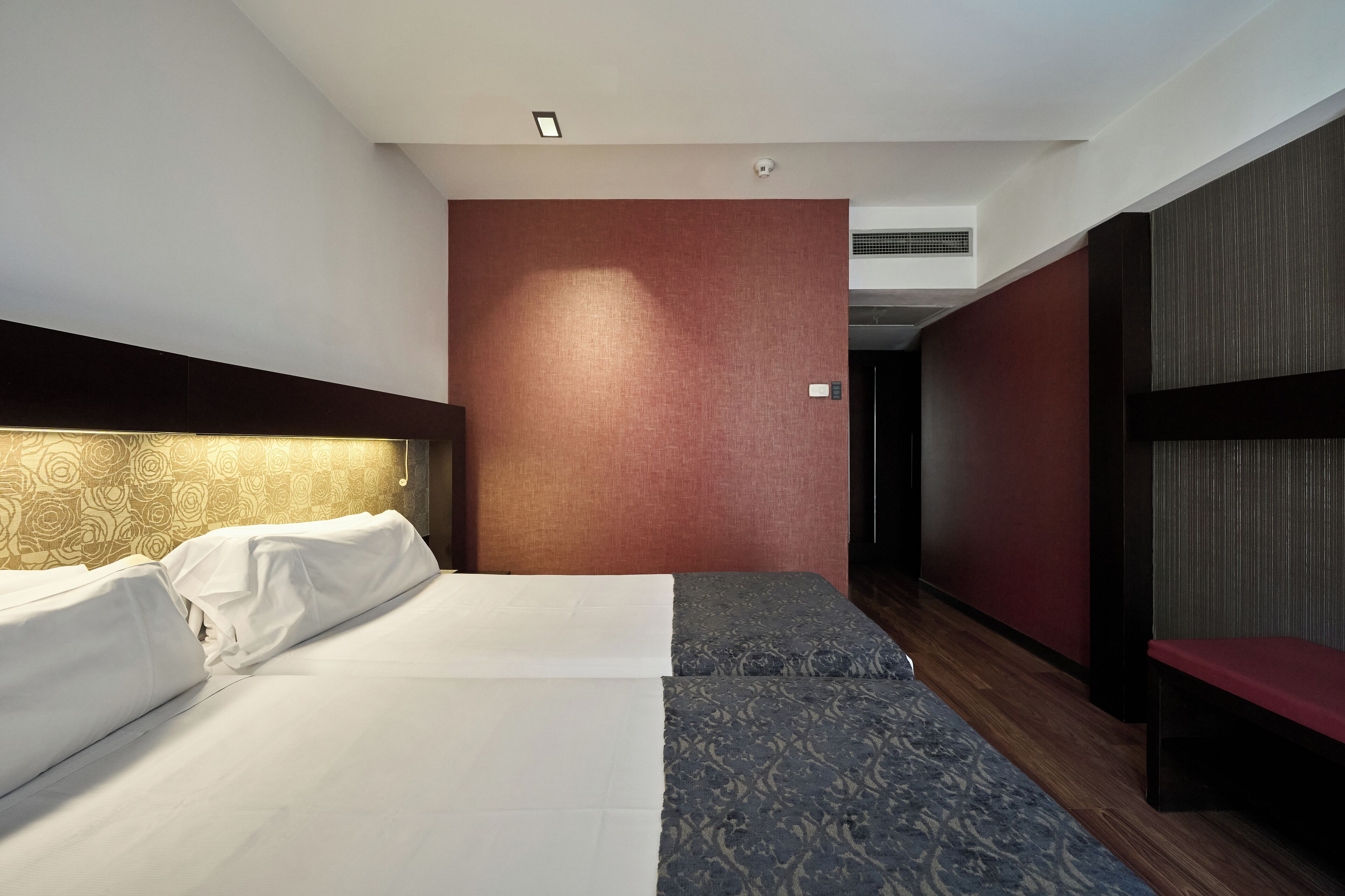 Ein Hotelzimmer mit Bett, Sitzbank, einem gemusterten Teppich und einer roten Akzentwand.