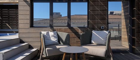 A modern outdoor seating area with a sofa and a table, wooden deck flooring, and large windows offering views of traditional buildings.