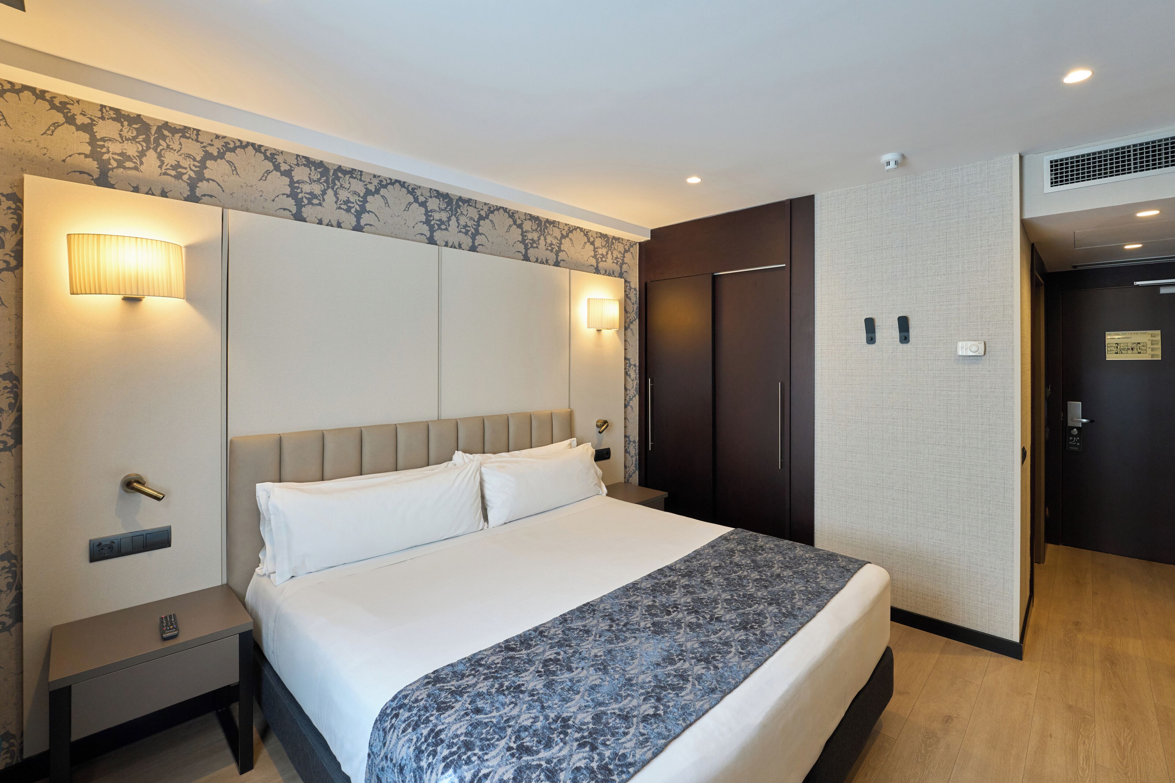 A hotel room with a bed, nightstands, wall sconces, a wardrobe, and a door with a sign.