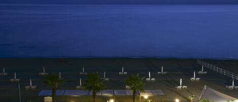 A beachside hotel with rows of sun loungers and umbrellas, a covered seating area, and a view of the ocean.