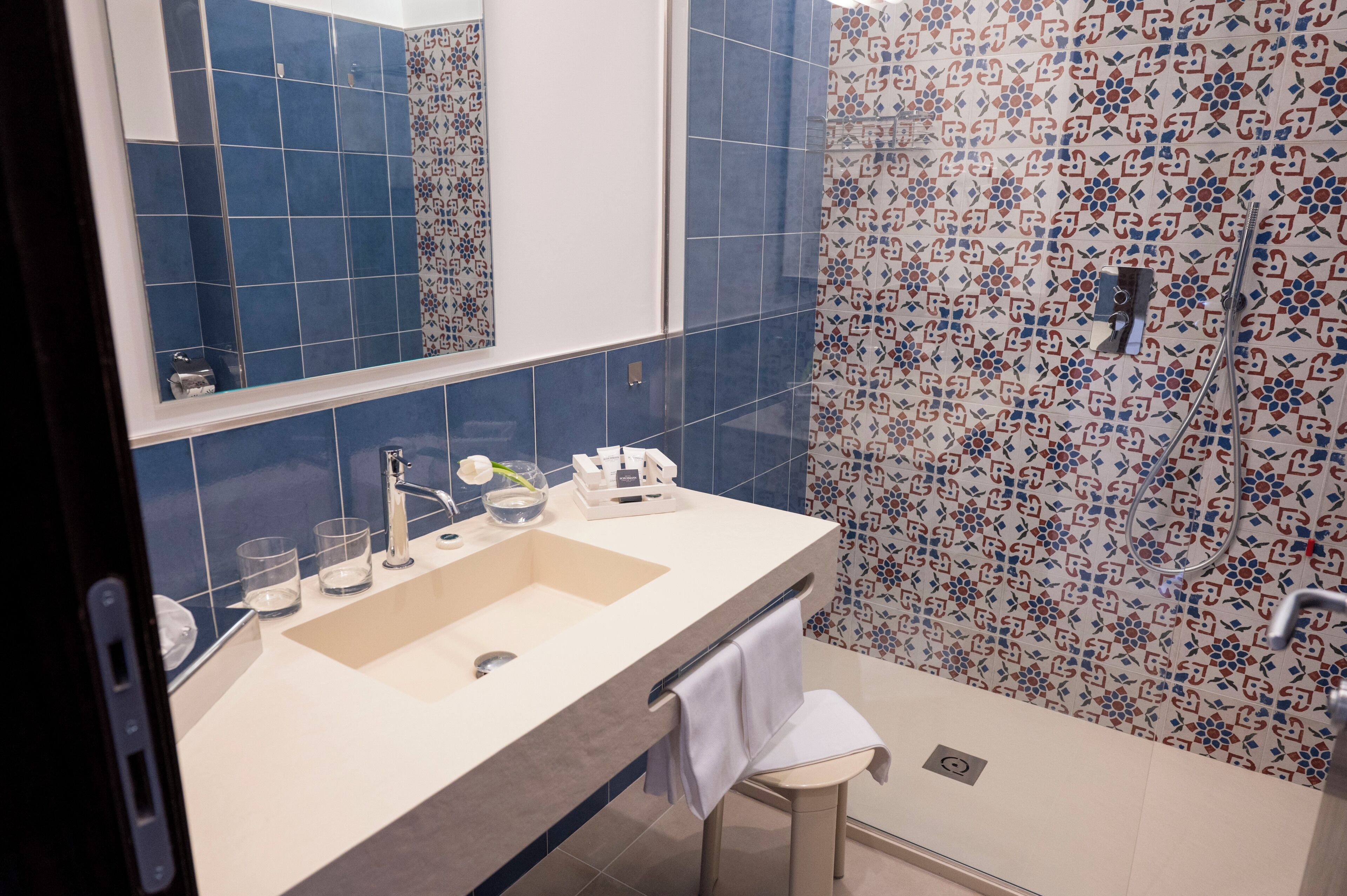 A bathroom with a large mirror, two sinks, a towel rack, and a shower area with patterned tiles.