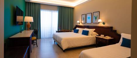 Habitación de hotel con dos camas, un escritorio y una silla. Hay cortinas, una lámpara y cuadros en la pared.