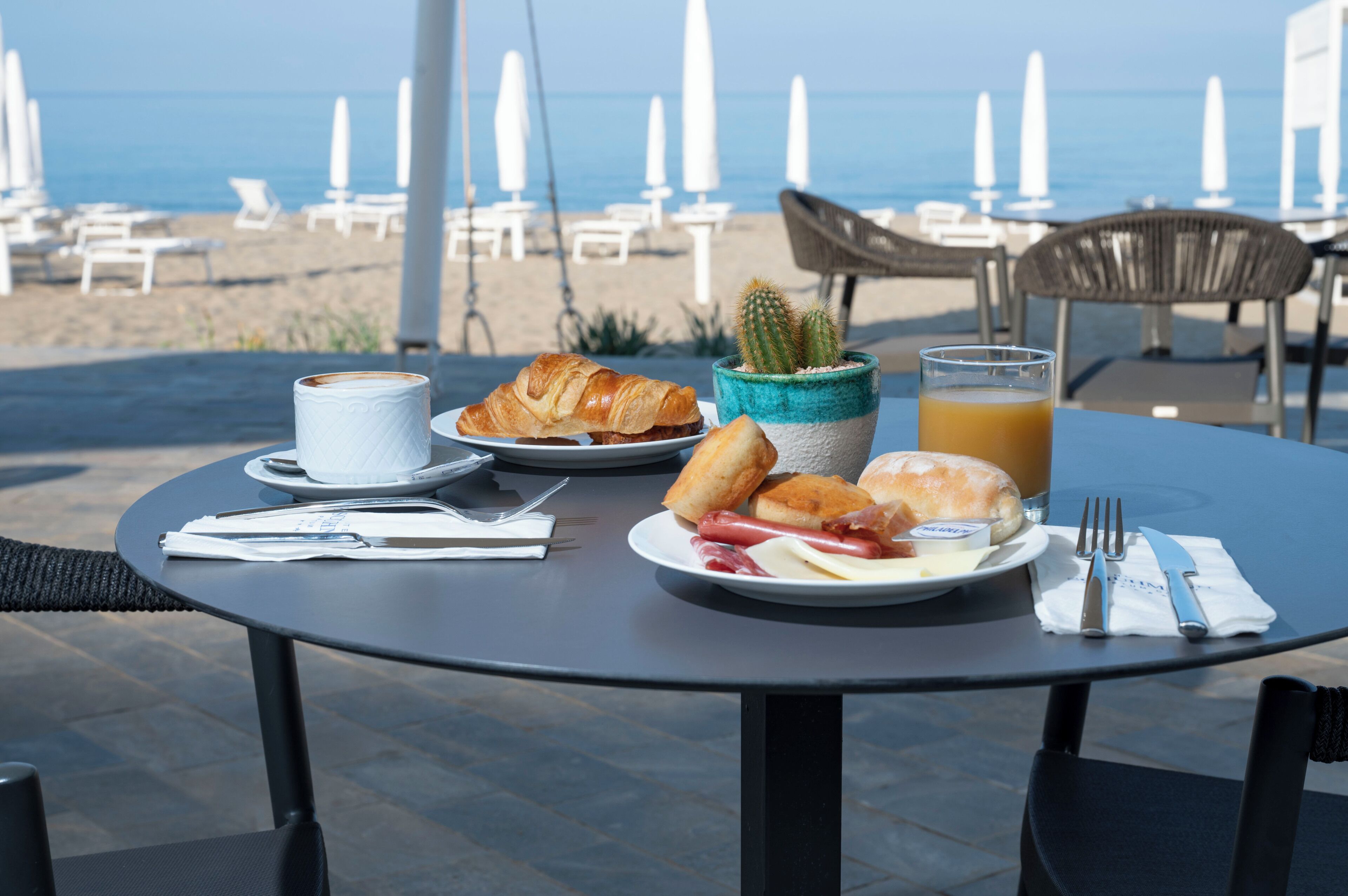 A table with croissants, a plate of sausages, a glass of orange juice, and a cup of coffee, set on a terrace with beach chairs and umbrellas in the background.