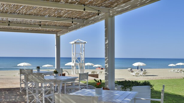 Un espace extérieur aménagé, abrit é et doté de tables et de chaises blanches, offrant une vue sur une plage parsemée de parasols et de chaises longues.
