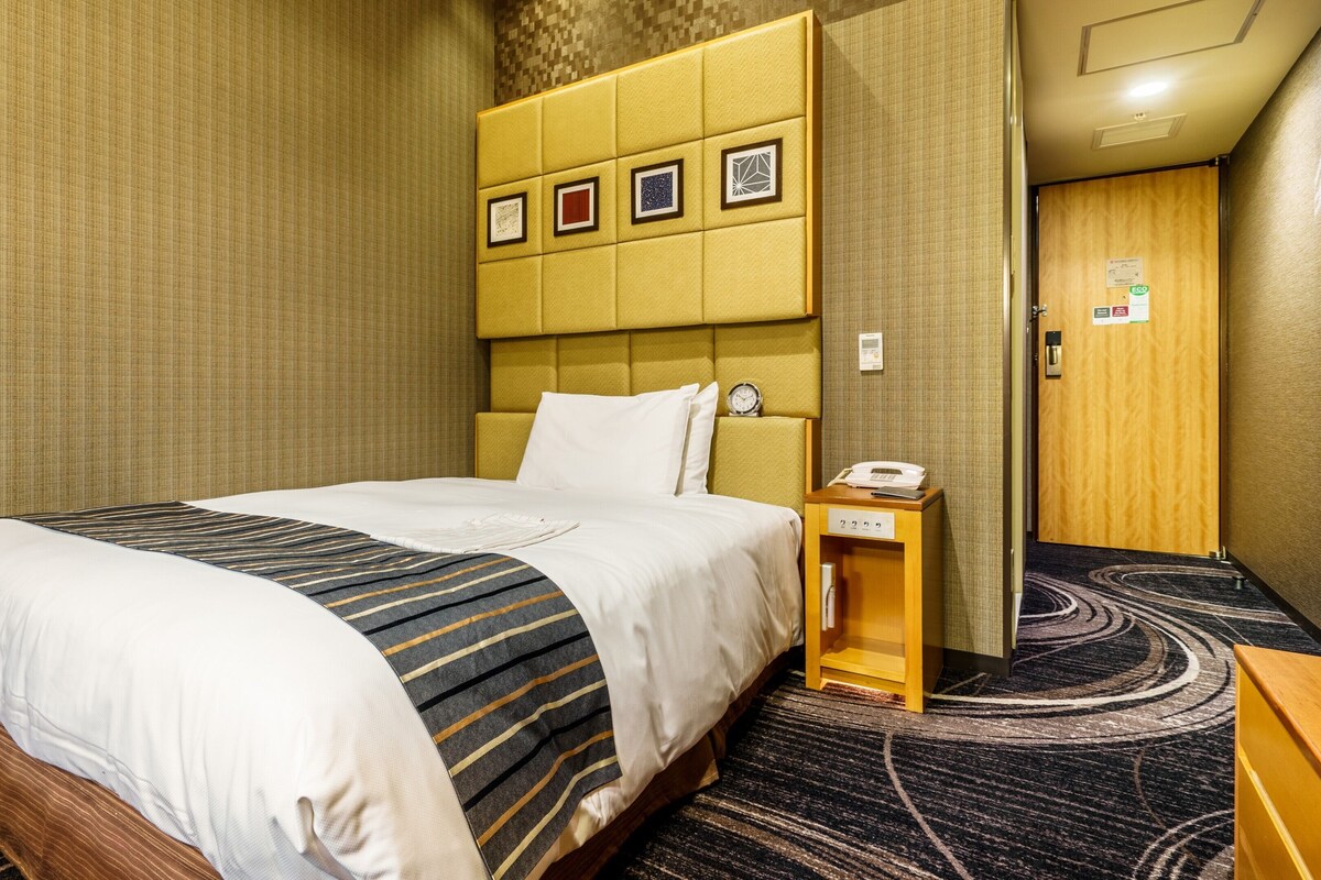 A hotel room with a bed, a bedside table with a phone, and a wall with framed pictures.