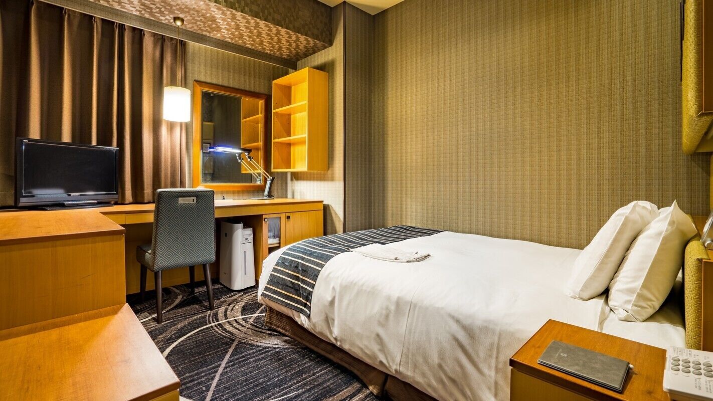 Ein Hotelzimmer mit einem Bett, einem Schreibtisch mit Computer, einem Stuhl, einer Lampe und einem kleinen Tisch mit einem Buch.