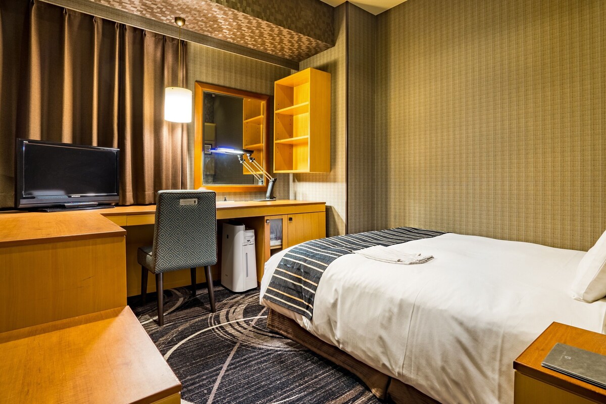 A hotel room with a bed, desk, chair, television, and a bedside table with a remote control.