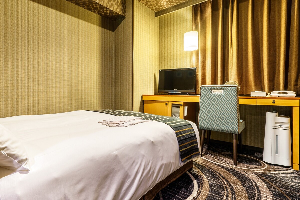 A hotel room with a bed, a desk with a computer, a chair, a telephone, and a waste bin.