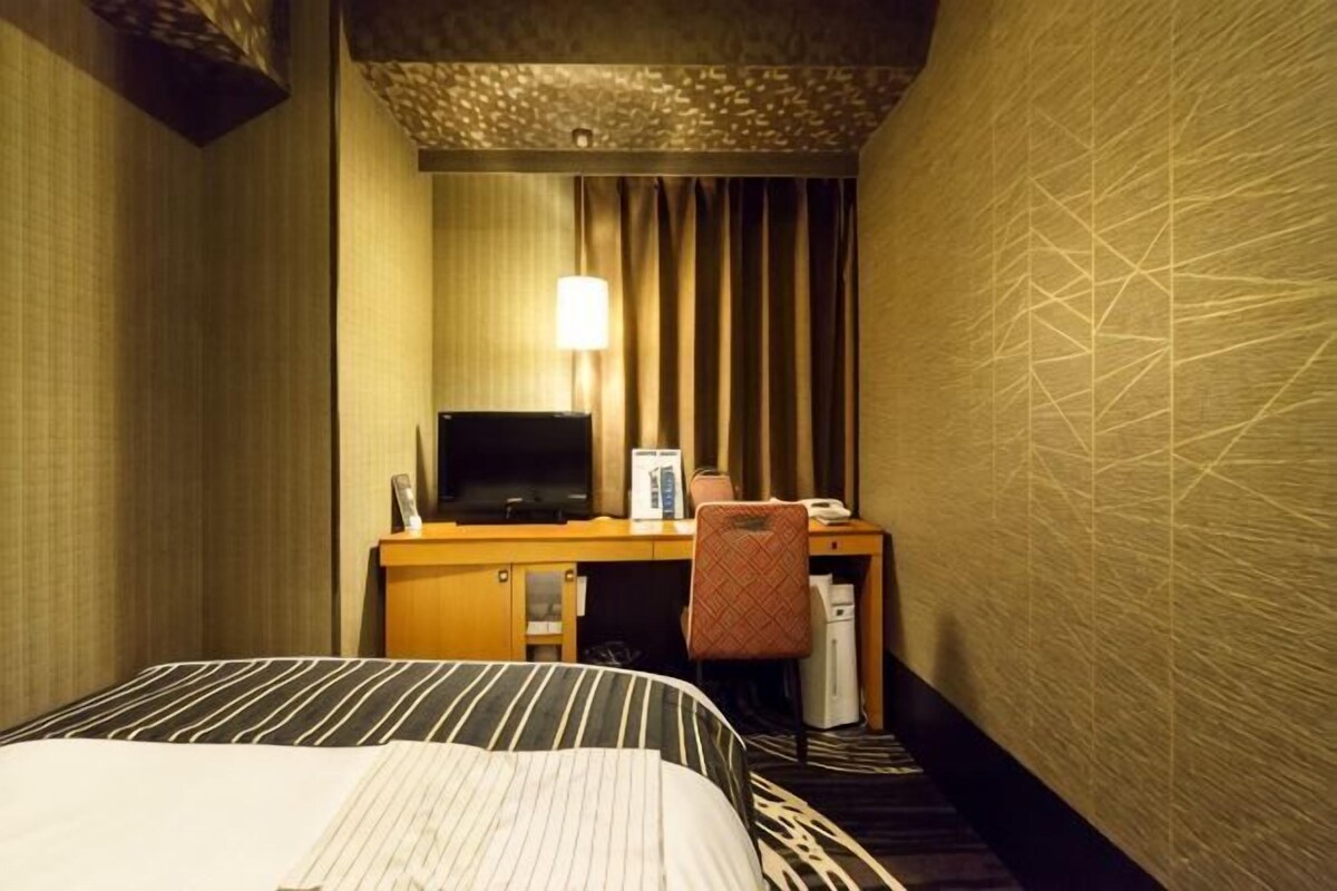 A hotel room with a bed, a desk with a television, a chair, and a lamp.
