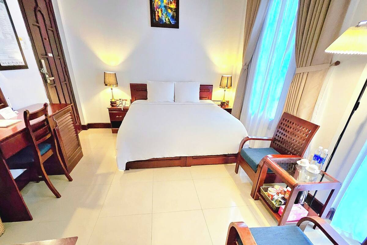 A hotel room with a large bed, a desk, a chair, and a small table with a lamp.