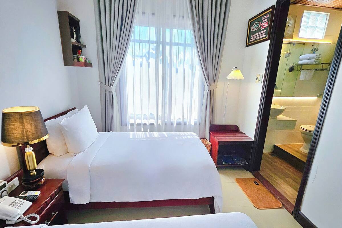 A hotel room with two beds, a desk with a lamp, a phone, and a bathroom visible through a glass door.