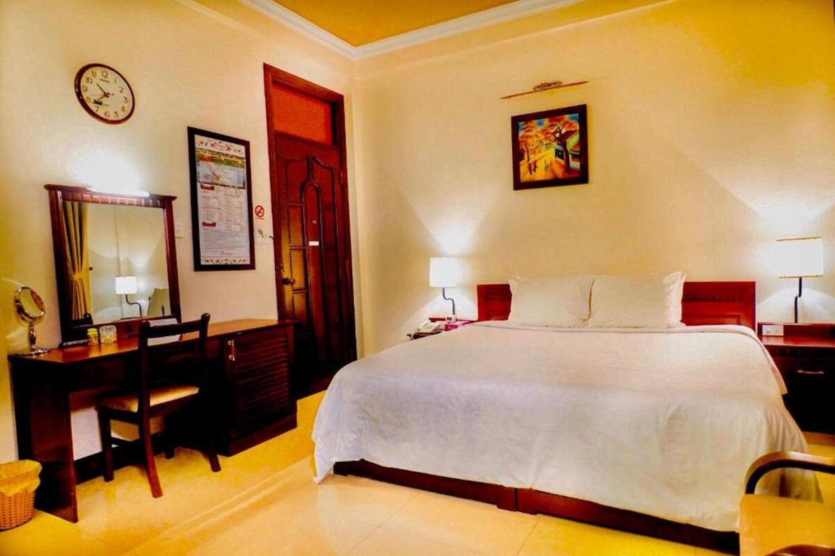 A hotel room with a bed, a desk with a chair, a mirror, a clock, and a door.