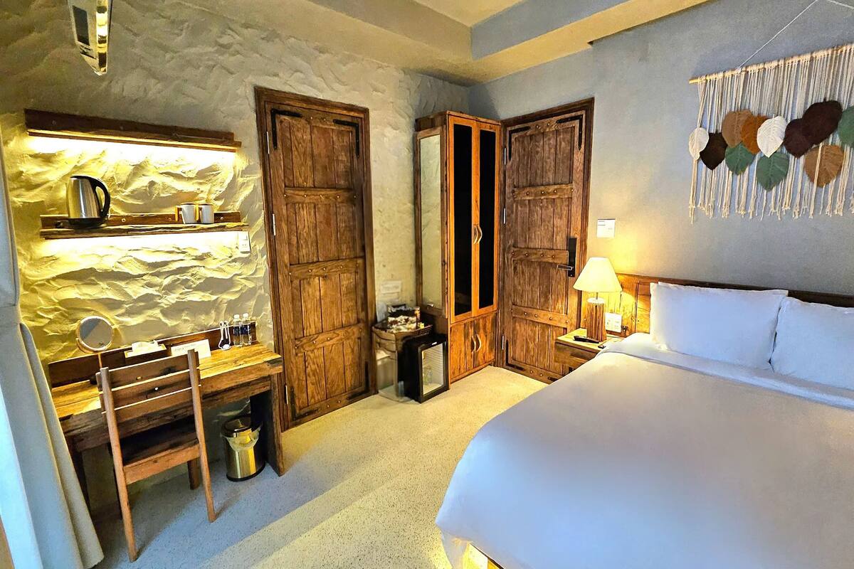 A bedroom with a bed, wooden wardrobe, a desk with a mirror, and a wall-mounted lamp.