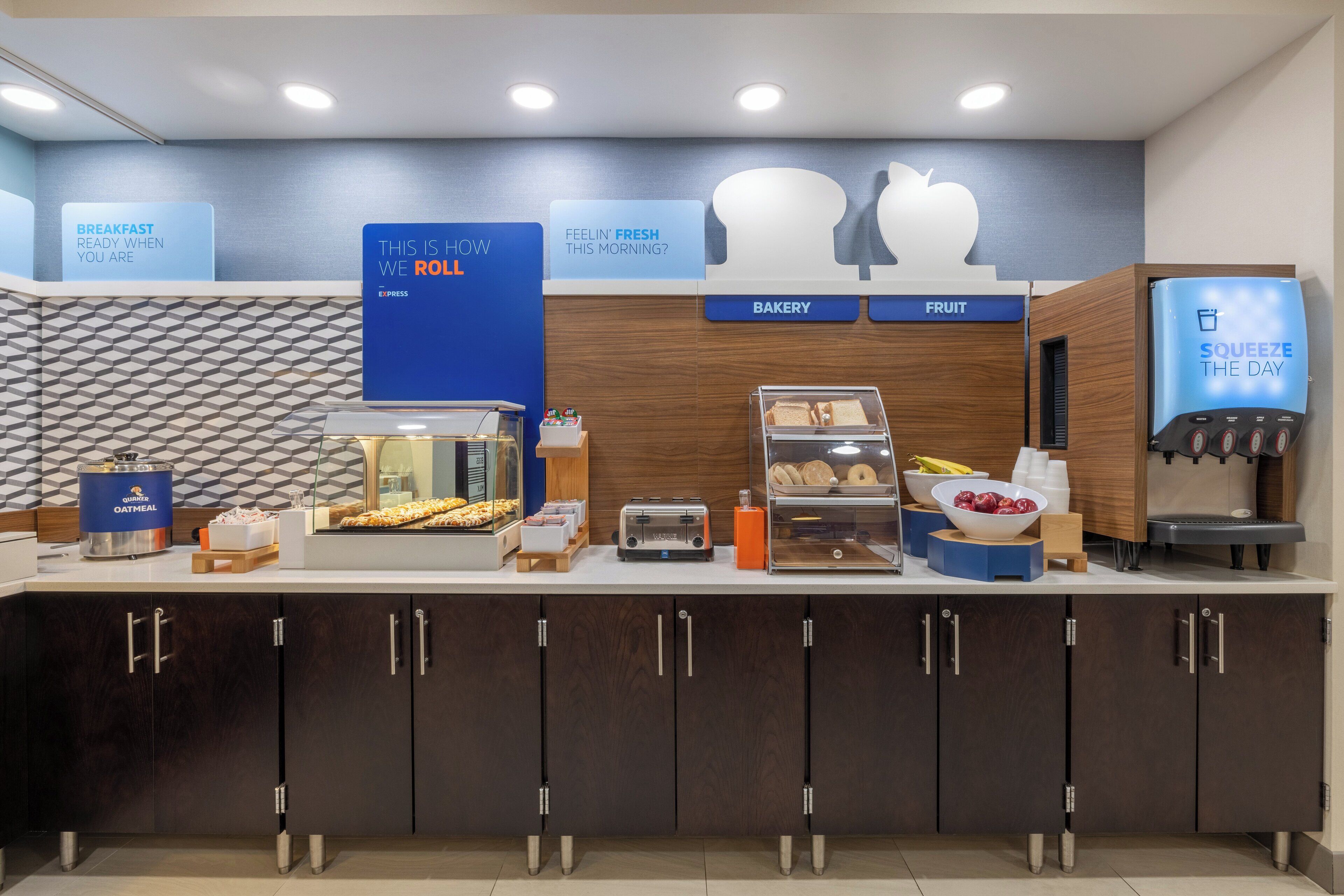 Un buffet pour le petit-déjeuner, avec des présentoirs étiquetés pour les produits de boulangerie, les fruits et un distributeur d’eau.