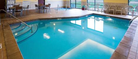 A swimming pool with a tiled deck, surrounded by chairs and tables, and a view of the outdoors through large windows.