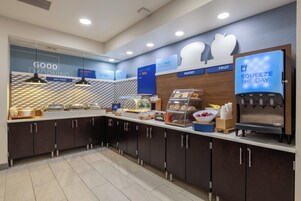 Un área de desayuno moderna con una barra que exhibe diversos alimentos, un dispensador de agua y un letrero que indica una sección de panadería.