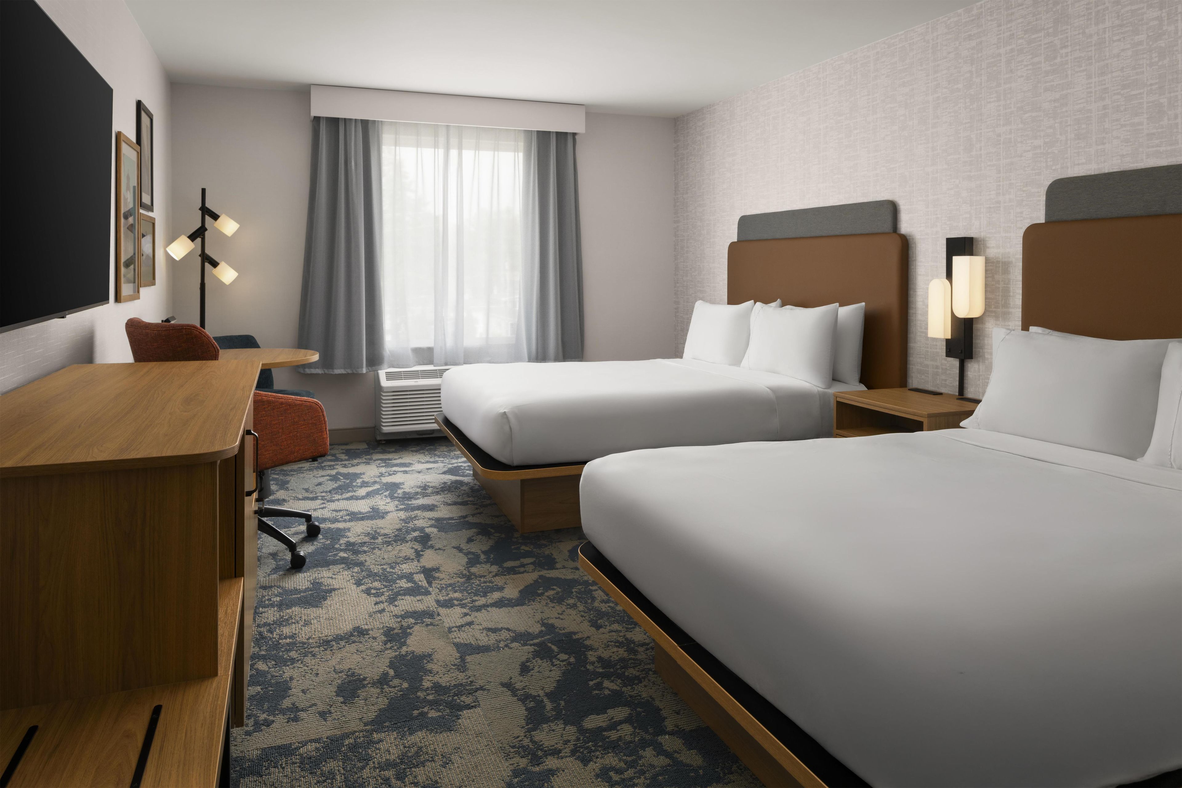 Habitación de hotel con dos camas, un escritorio, una silla, un ventanal con cortinas y una lámpara de pared.