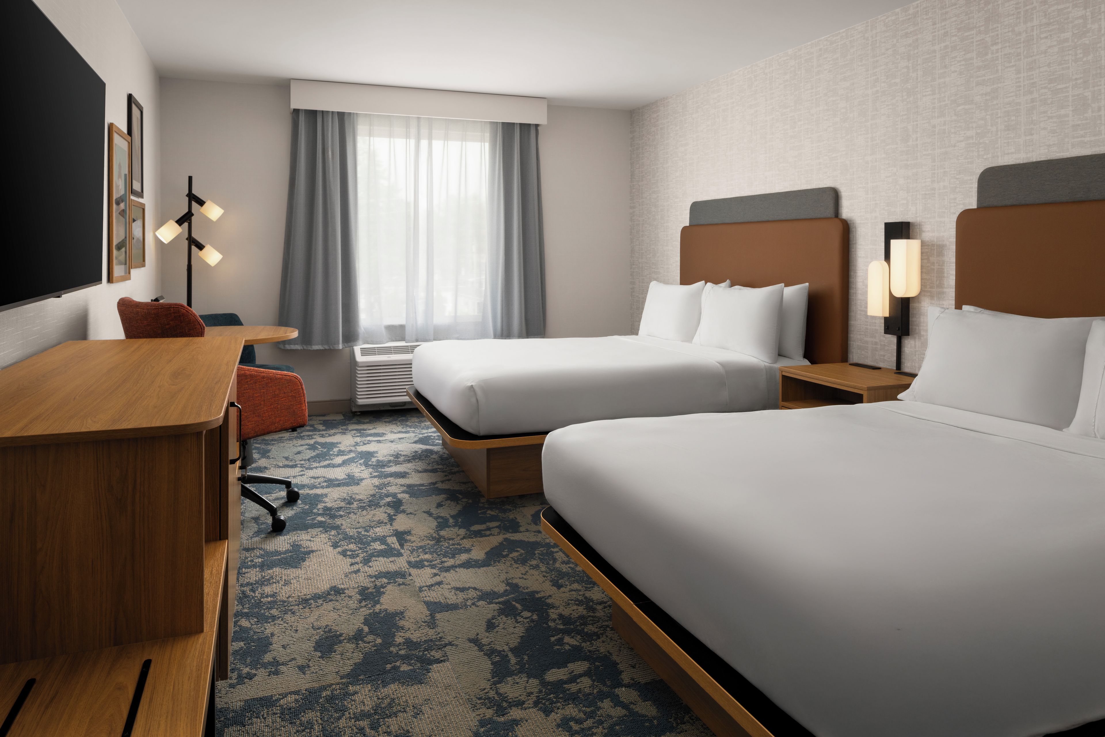 Une chambre d’hôtel avec deux lits, un bureau, une chaise, une fenêtre avec des rideaux et une lampe fixée au mur.