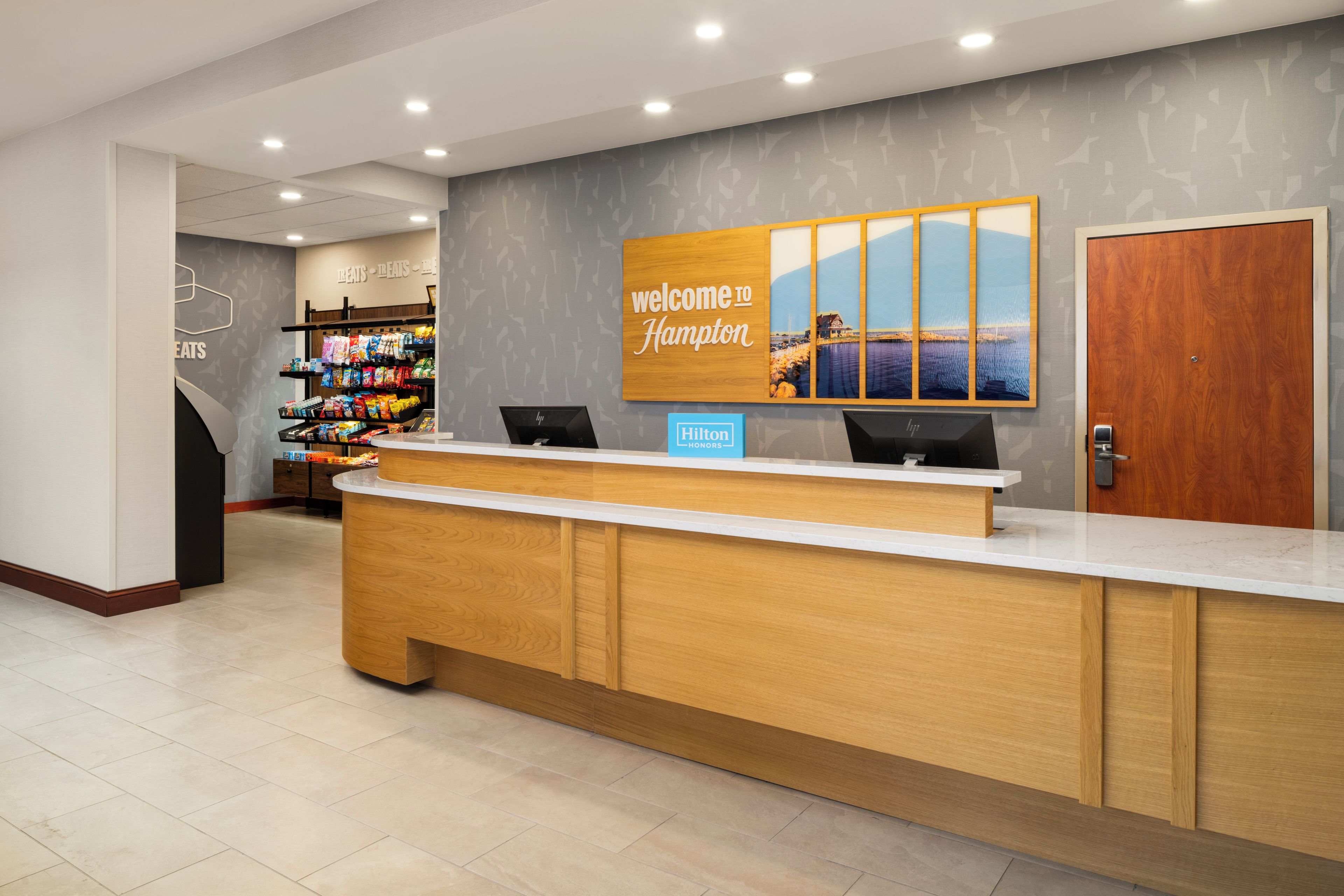 Ein Hotelrezeptionstresen mit einem 'Welcome to Hampton'-Schild, einem Computer und einer Snackauslage.