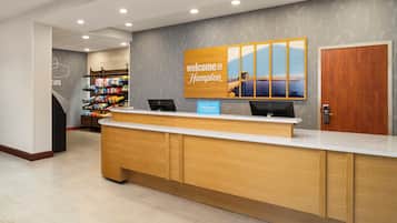 Un mostrador de recepción de hotel con un cartel que dice 'Welcome to Hampton', una computadora y una exhibición de refrigerios.