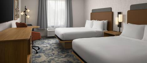 A hotel room with two beds, a desk, a chair, a window with curtains, and a wall-mounted lamp.