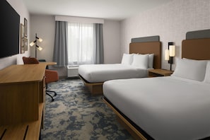 A hotel room with two beds, a desk, a chair, a window with curtains, and a wall-mounted lamp.