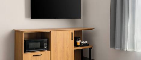 A room with a flat-screen TV mounted on the wall, a wooden cabinet with a microwave, and a small table with a coffee maker and a cup.