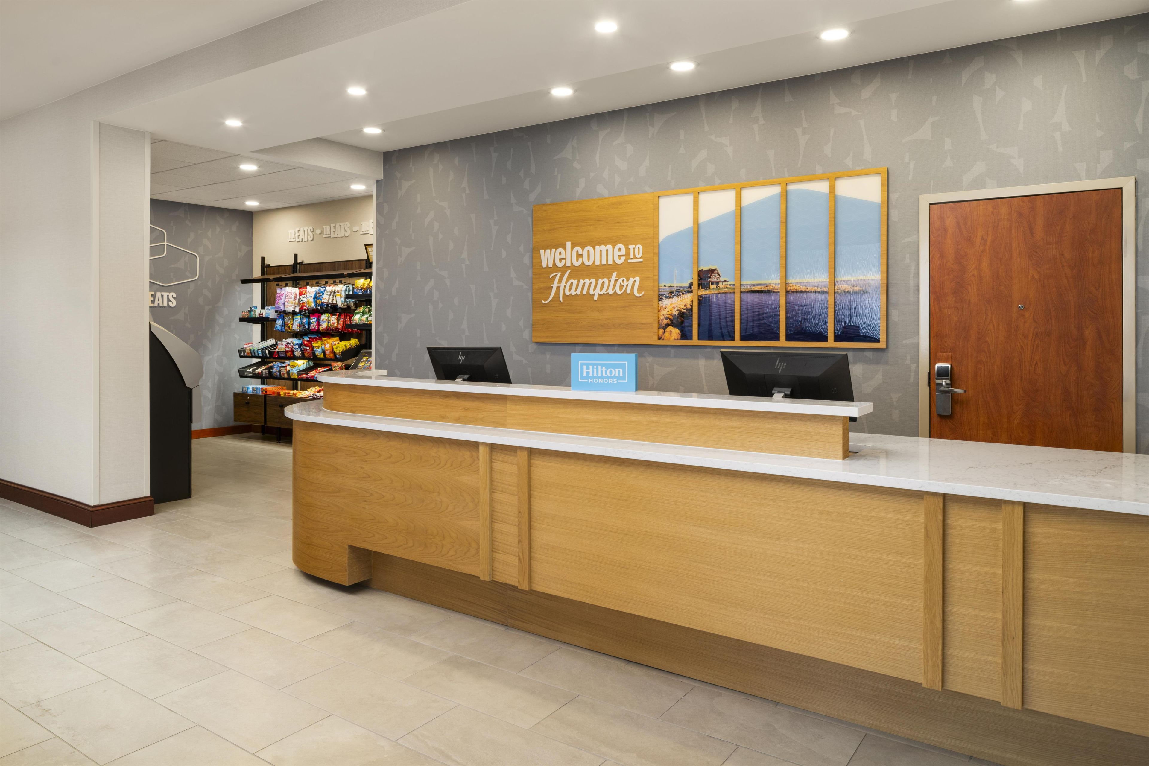 A hotel reception area with a wooden reception desk, a 'Welcome to Hampton' sign, a view of the sea, and a snack bar in the background.