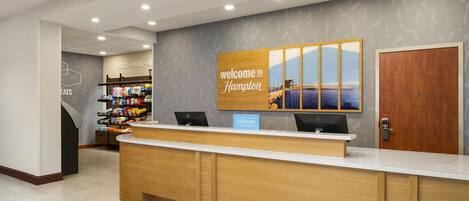 A hotel reception area with a wooden reception desk, a 'Welcome to Hampton' sign, a view of the sea, and a snack bar in the background.