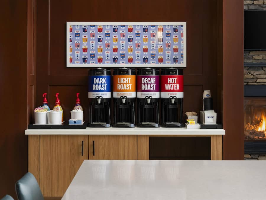 A coffee station with four dispensers labeled DARK ROAST, LIGHT ROAST, DECAF ROAST, and HOT WATER, and a wall-mounted coffee cup display above.