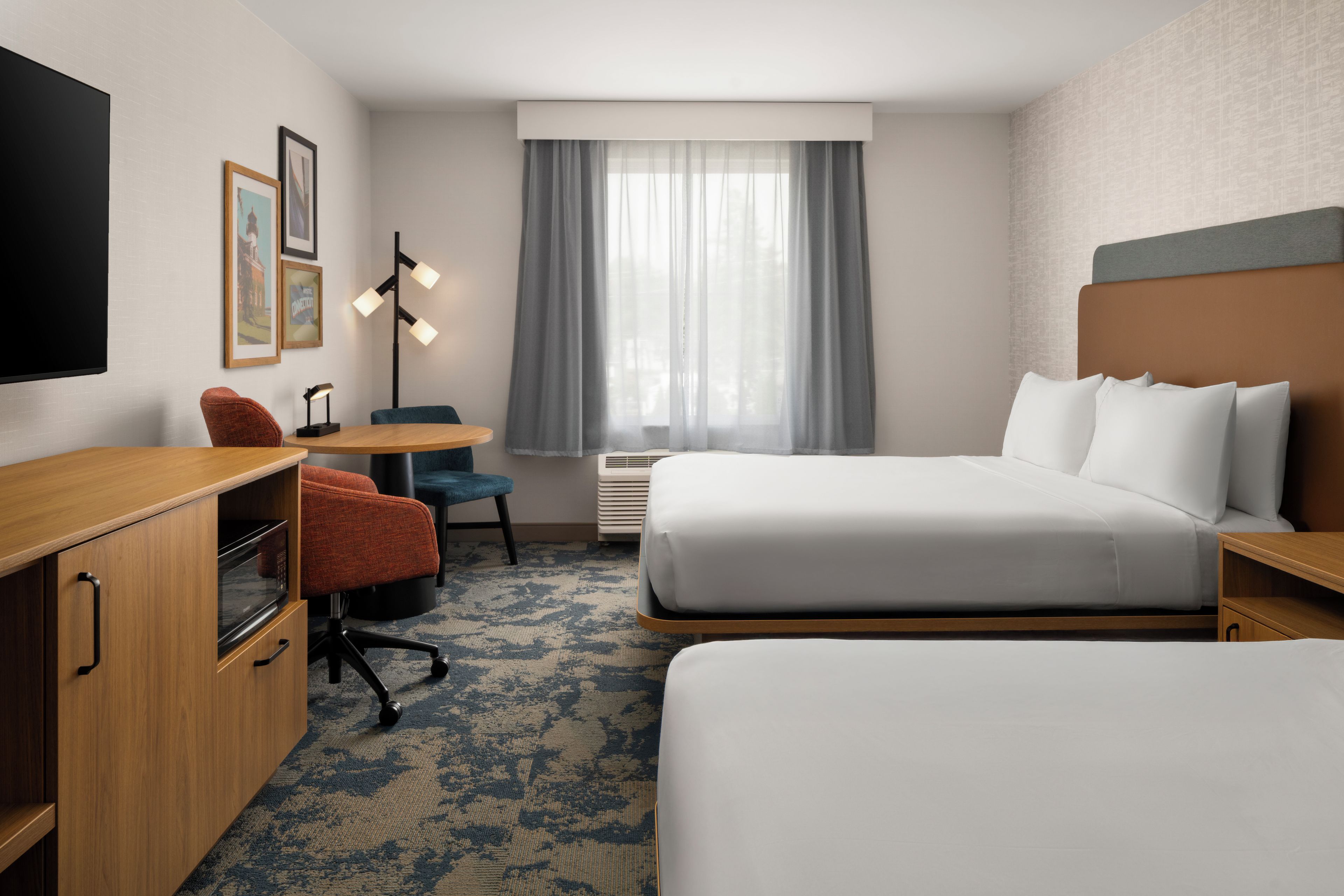 Quarto de hotel com cama, escrivaninha, cadeira, televisão e janela com cortinas.