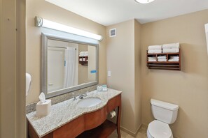 A bathroom with a sink, mirror, and toilet. There is a towel rack above the toilet and a shelf with towels.
