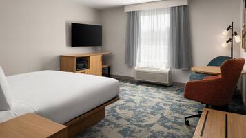 Habitación de hotel con cama, escritorio, silla, televisor, equipo de aire acondicionado y ventana con cortinas.