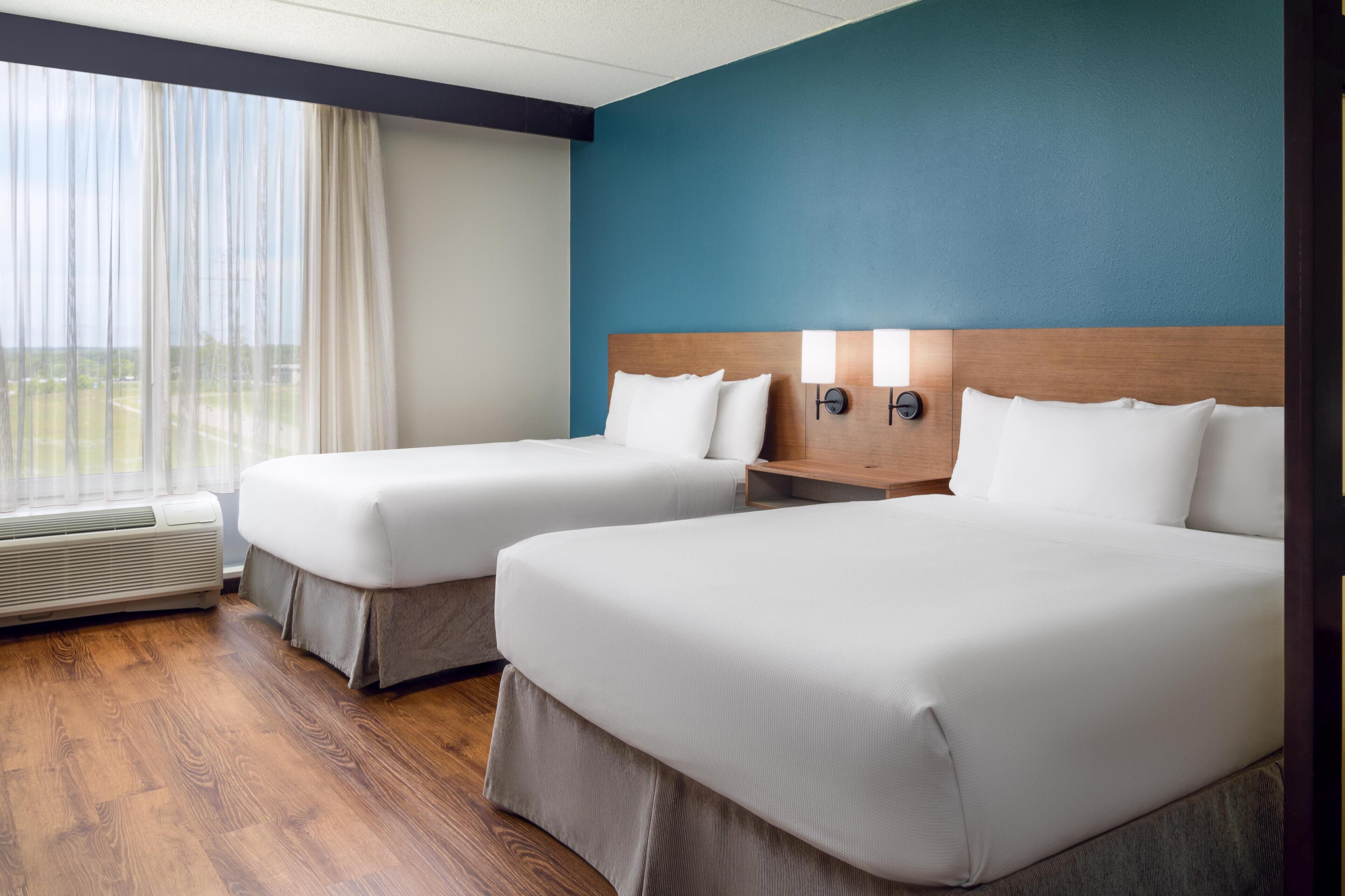 A hotel room with two beds, a wooden headboard, a nightstand with lamps, and a large window with curtains.
