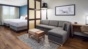Ein Hotelzimmer mit zwei Betten, einem grauen Sofa, einem Holztisch, einem Schreibtisch und einem gerahmten Bild an der Wand.