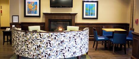 A lobby with a curved reception desk, a flat-screen TV mounted above it, and a fireplace. There are framed artworks on the walls and a seating area with chairs and a table.