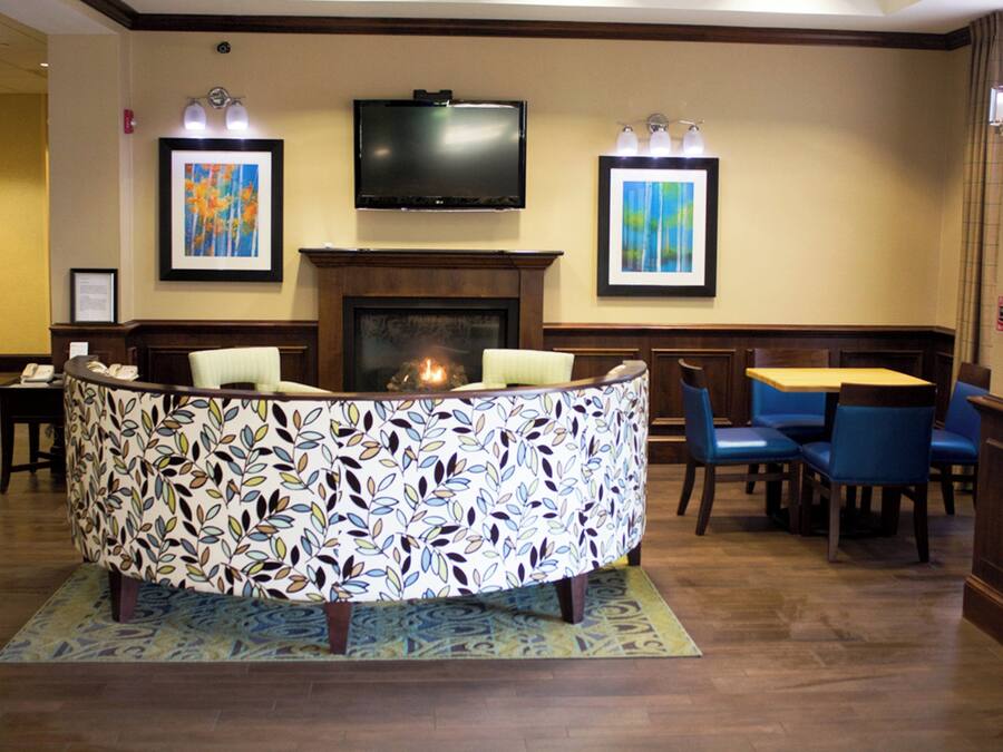 A lobby with a curved reception desk, a flat-screen TV mounted above it, and a fireplace. There are framed artworks on the walls and a seating area with chairs and a table.