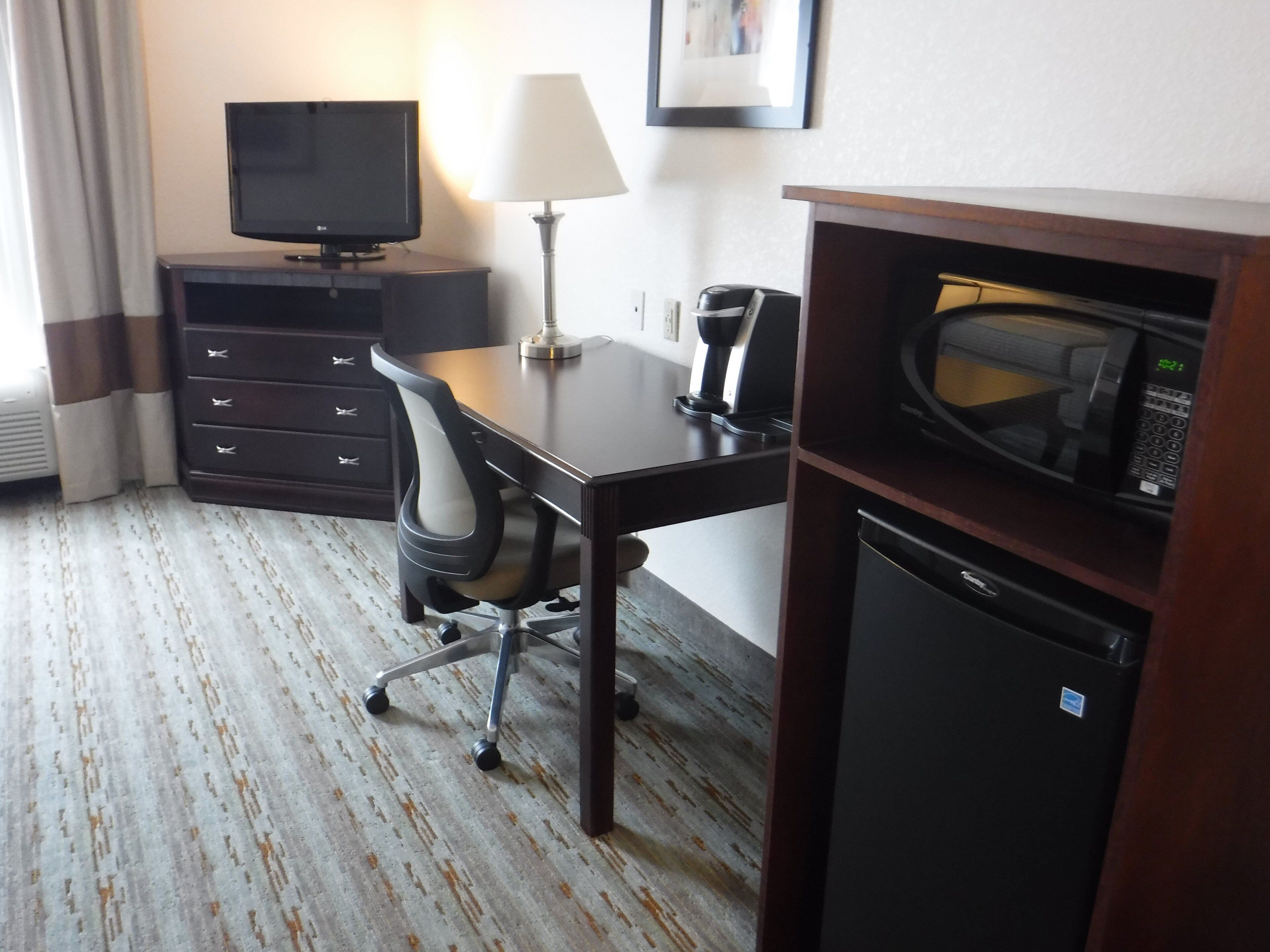 A hotel room with a desk, a chair, a television, a lamp, a microwave, and a mini fridge.