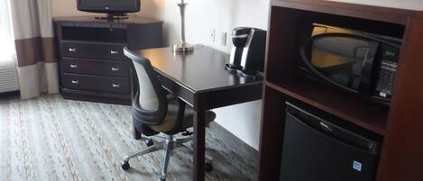 A hotel room with a desk, a chair, a television, a lamp, a microwave, and a mini fridge.