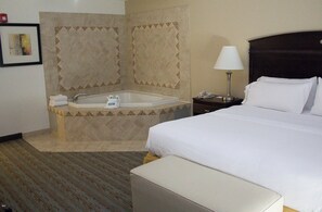 A hotel room with a large bed, a nightstand with a lamp, a bathroom with a bathtub, and a framed picture on the wall.