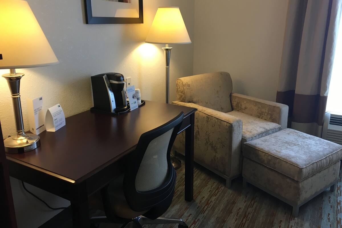 A hotel room with a desk, two lamps, a coffee maker, a chair, and an ottoman.