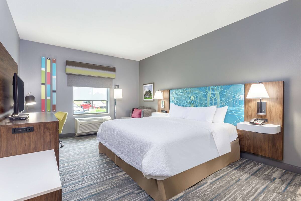 A modern hotel room with a large bed, a desk, a chair, a TV, a window with a view, and a map artwork on the wall.