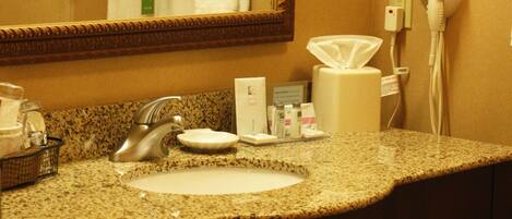 Un baño de hotel con encimera de granito, un lavabo, un espejo, un perchero con toallas dobladas, un secador de pelo y un cubo de basura.