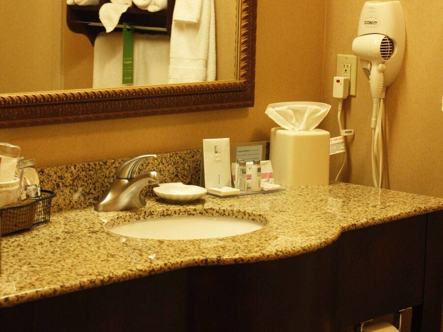 Une salle de bain d’hôtel avec un comptoir en granit, un lavabo, un miroir, un porte-serviettes avec des serviettes pliées, un sèche-cheveux et une poubelle.