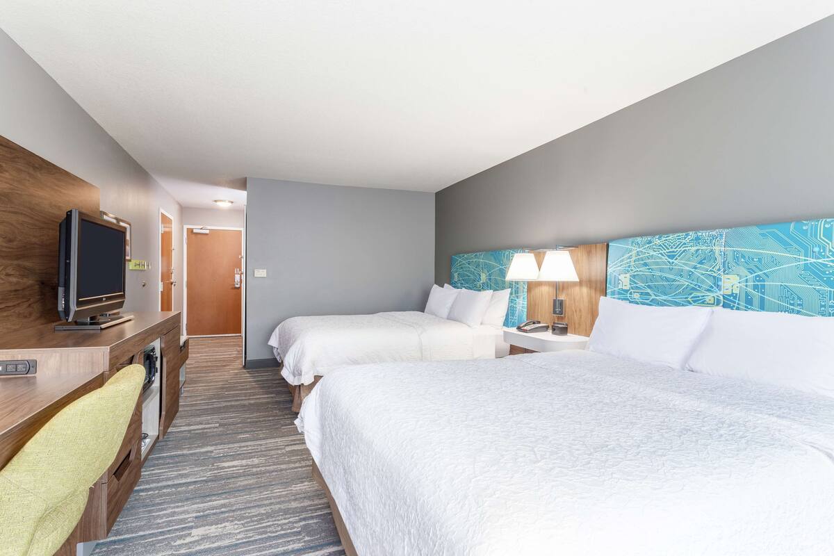 A hotel room with two beds, a desk with a television, a lamp, and a headboard with a circuit board design.