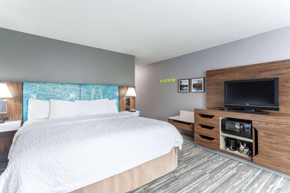 A hotel room with a large bed, a flat-screen TV, a wooden cabinet with a microwave, and a desk with a lamp.