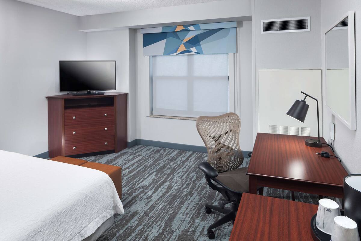 Ein Hotelzimmer mit einem Bett, einem Schreibtisch mit Lampe, einem Sessel, einem Fernseher auf einer Holzkommode und einem Fenster mit Jalousien.